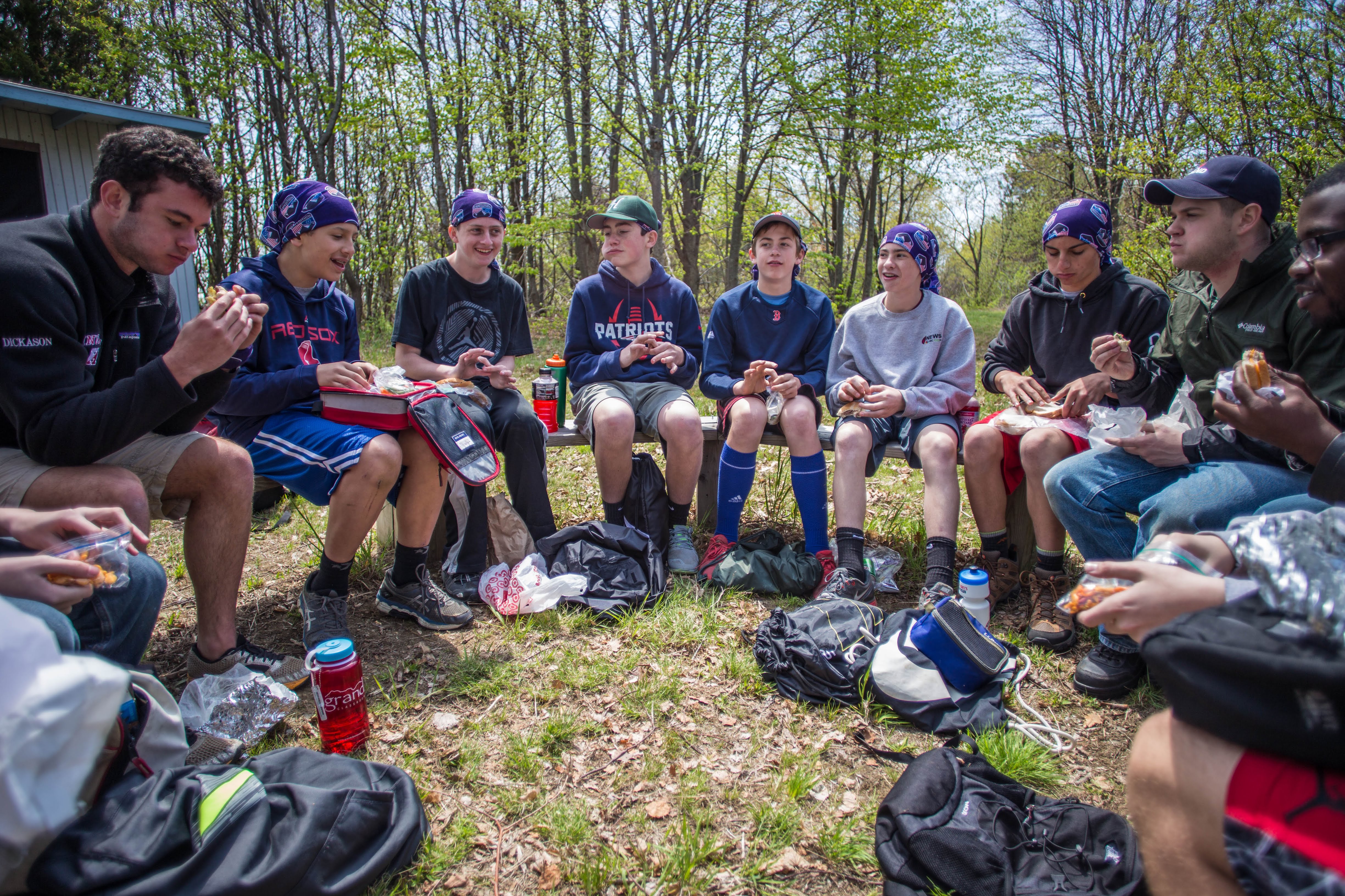 The image shows a group of young people sitting in a circle outdoors, likely during a break. They are eating, and some have food containers. They are surrounded by trees, suggesting a park or outdoor setting. The atmosphere seems relaxed and social.